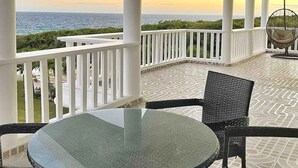 A covered outdoor seating area with a glass table and wicker chairs, overlooking a body of water at sunset.