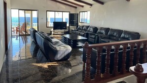 A spacious living room with a black leather sofa, a television, and a large window offering a view of the ocean.