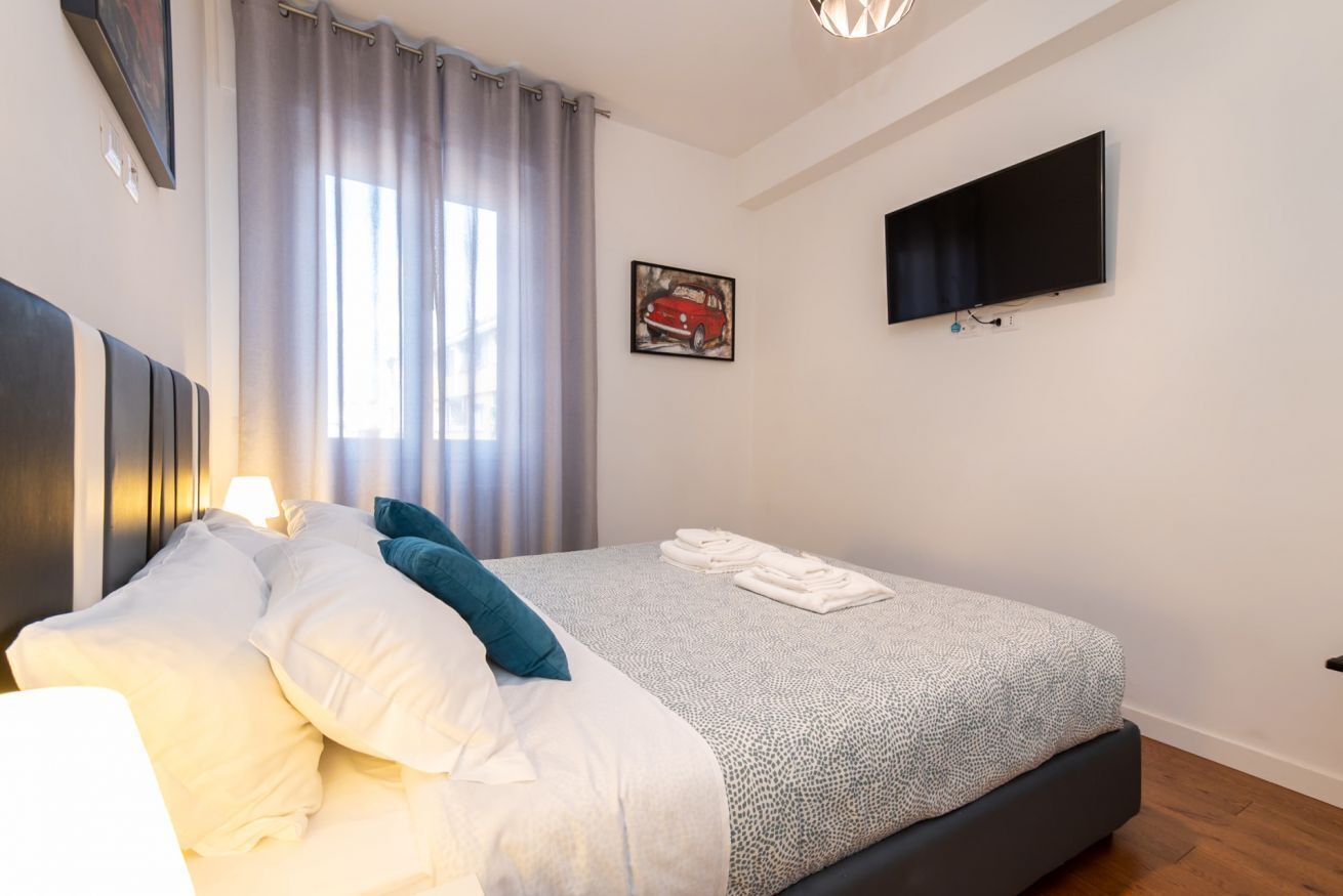 A neatly made bed with white and teal pillows, a grey patterned bedspread, and folded towels on the side. A flat-screen TV is mounted on the wall above the bed.