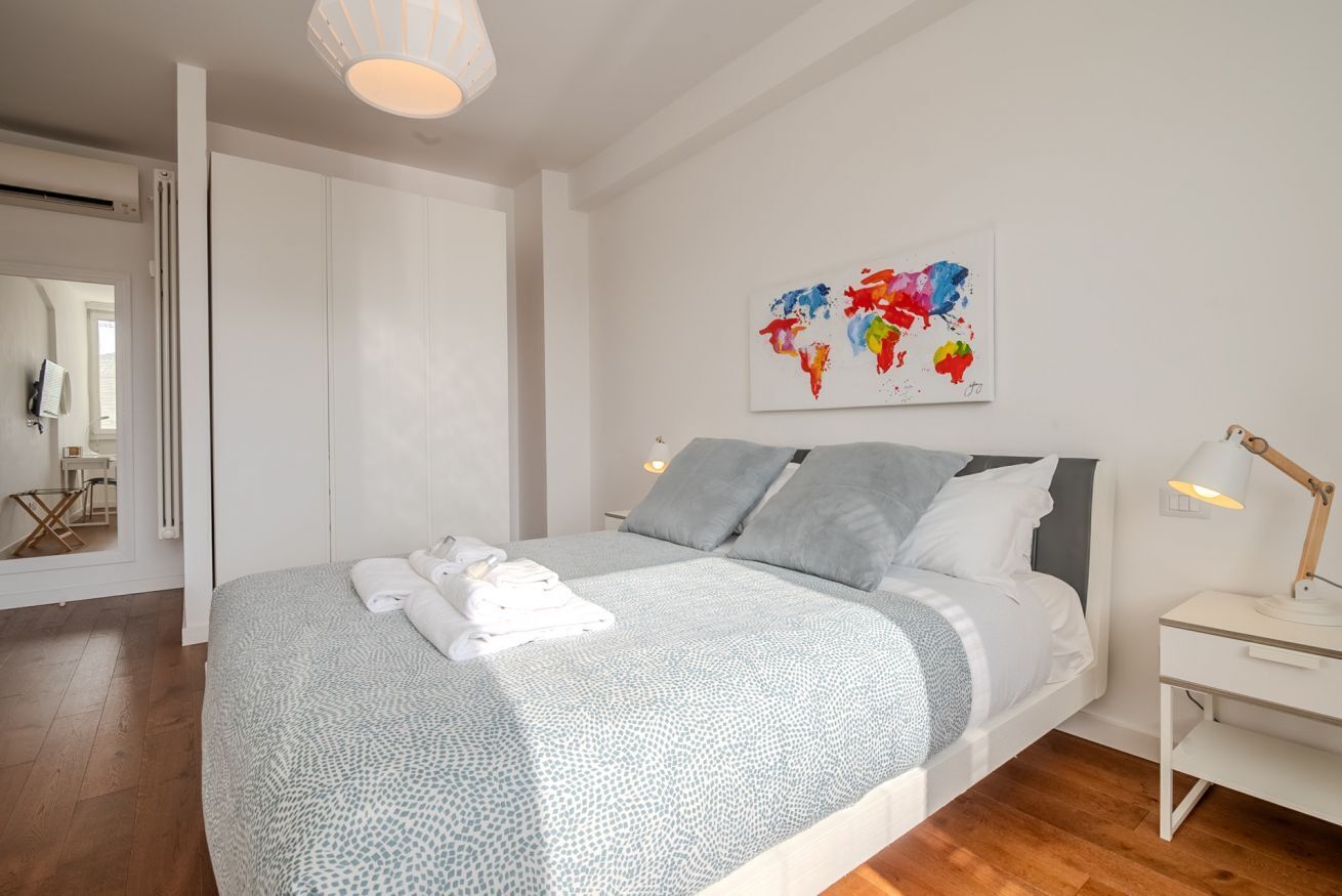 A modern bedroom with a large bed, white walls, a world map artwork, and a wooden floor.