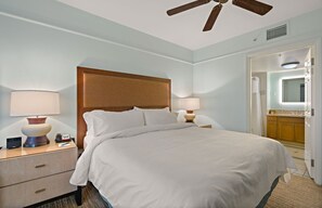 Ein Hotelzimmer mit einem großen Bett, zwei Nachttischen mit Lampen, einem Holzkopfstück, einem Deckenventilator und einem durchgegangenen Türrahmen erkennbaren Badezimmer.