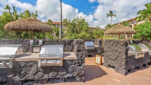 An outdoor barbecue area with stone walls and thatched umbrellas, surrounded by lush greenery and a clear blue sky. - Marriott's Waiohai Beach Club 2BR Islandview Villa (Koloa)
