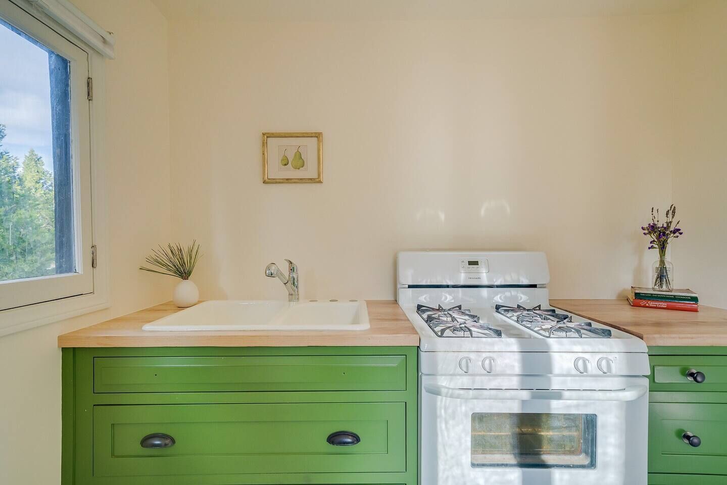Una cocina con estufa blanca, armarios verdes, un lavabo blanco y una ventana con vistas a árboles.