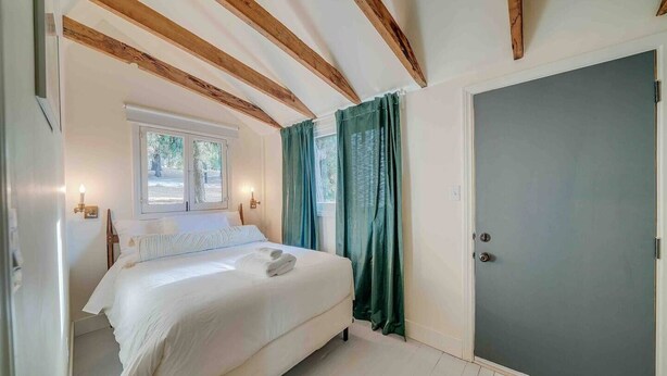 A bedroom with a bed, wooden beams, a window, curtains, and a door.