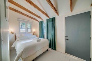 A bedroom with a bed, wooden beams, a window, curtains, and a door.