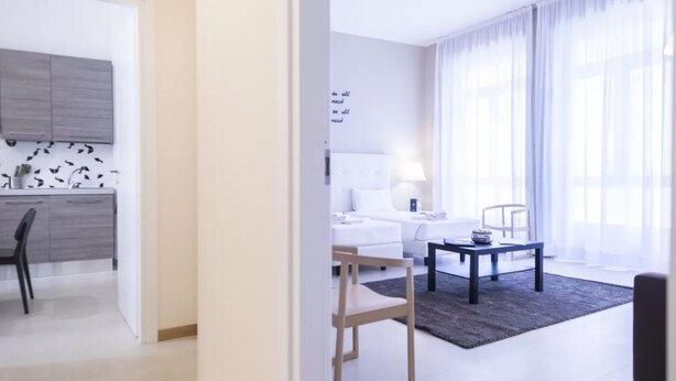 Une chambre d’hôtel moderne dotée d’une kitchenette, d’un coin repas et d’une grande fenêtre avec des rideaux légers.