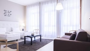 Un salon moderne avec un canapé blanc, un canapé marron, une table basse et un fauteuil. Une grande fenêtre est agrémentée de rideaux légers et une lampe est fixée au mur.
