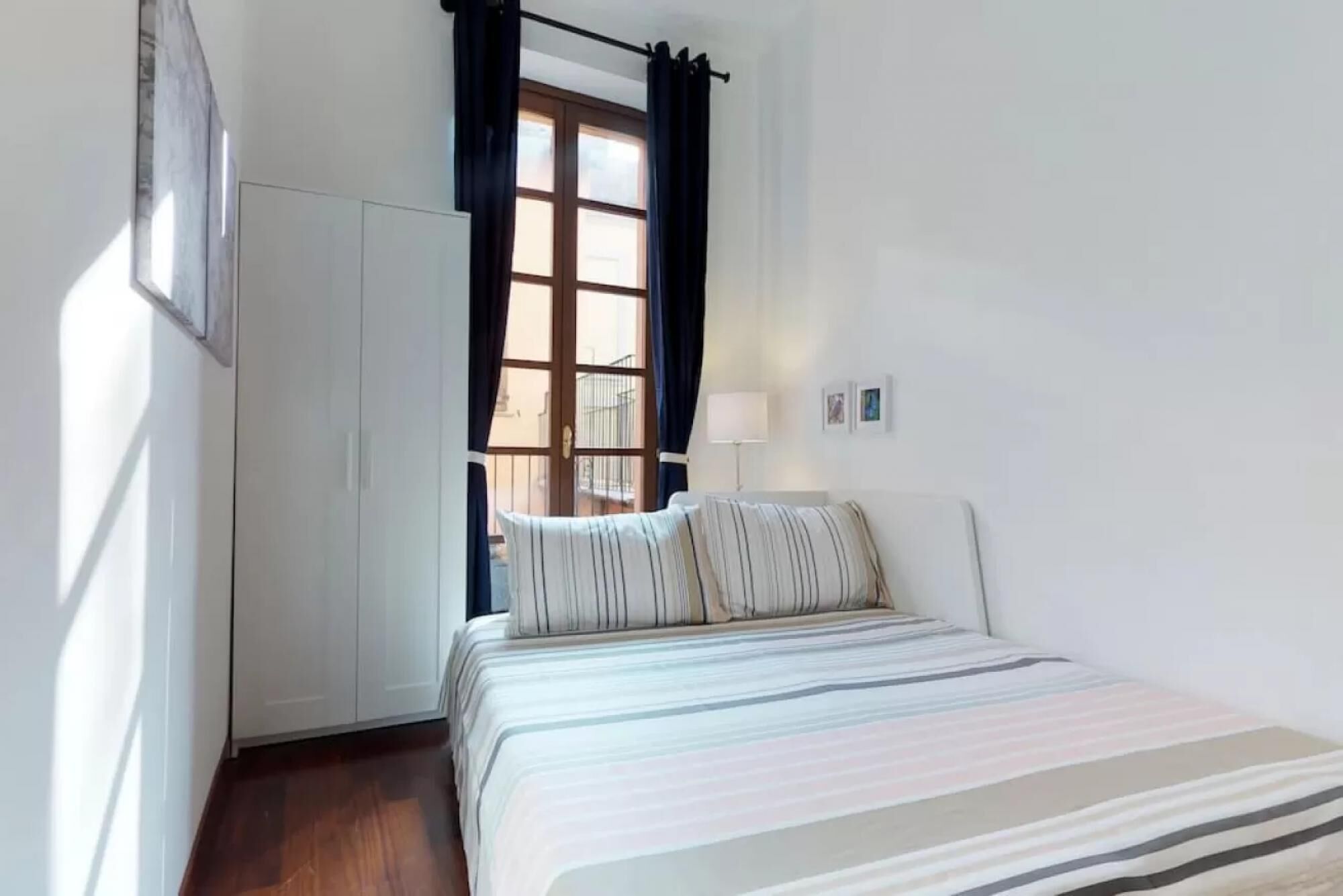 A bedroom with a bed, a wardrobe, a window with curtains, and a wall-mounted lamp.