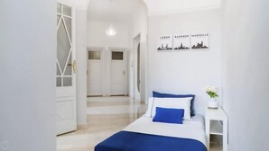 Une chambre d’hôtel avec un lit, une table de chevet, un mur décoré d’une œuvre d’art représentant un paysage urbain, et un couloir avec des portes.