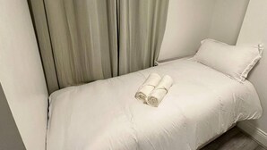 A single bed with a white duvet, a pair of rolled towels, and a grey curtain.