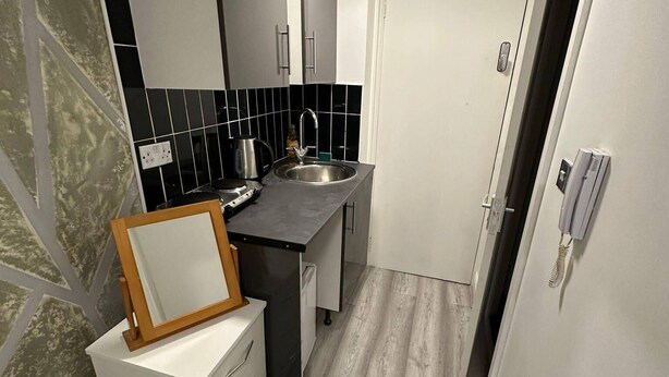 A compact kitchen with a sink, countertop, and cabinets. There is a wooden frame leaning against a white cabinet. The floor is wooden, and the walls are white with black tiles.