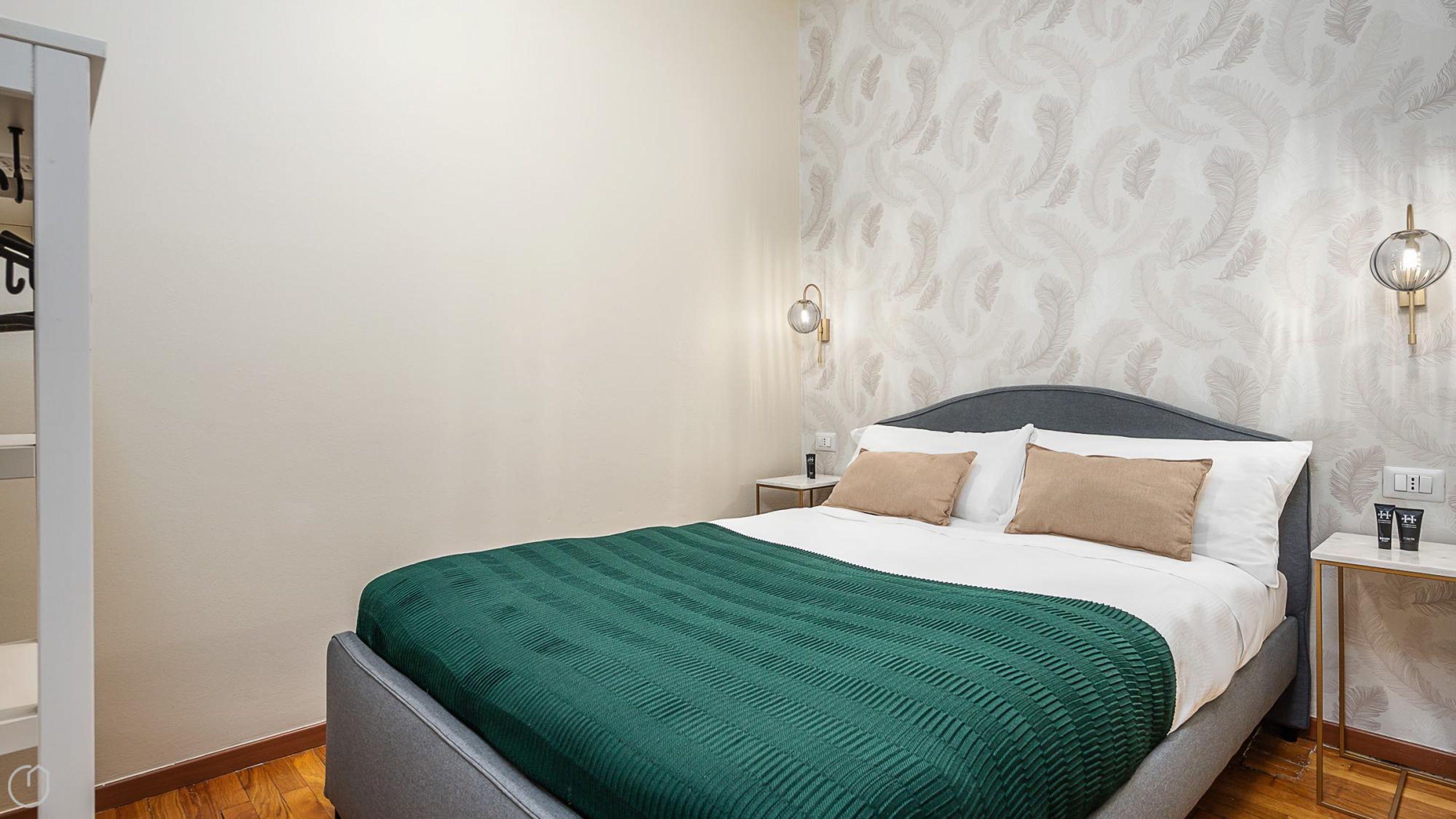 A neatly made bed with a green blanket and two pillows, a bedside table with a lamp, and a wall with a leaf pattern.