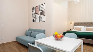 Une chambre d’hôtel moderne avec un canapé bleu, une table blanche sur laquelle se trouve un panier de fruits, un lit recouvert d’une couverture verte et un lustre.