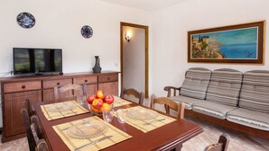 Een eethoek met een houten tafel voor vier personen, een televisie op een kast, en een bank met een schilderij van een kustlandschap.