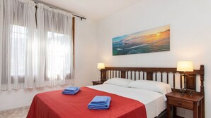 A hotel room with a bed, two bedside tables with lamps, a painting of a sunset, and a window with sheer curtains.