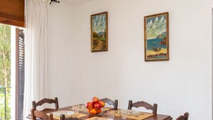 A dining table with a fruit bowl, set with glasses and cutlery, in a room with two paintings on the wall and a view through a window.