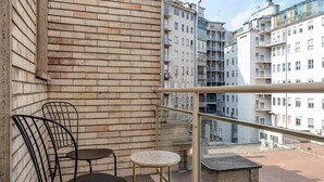 Un balcón con pared de ladrillo, dos sillas de metal, una mesa redonda pequeña y barandilla de vidrio con vistas a una zona residencial con edificios de varios pisos.
