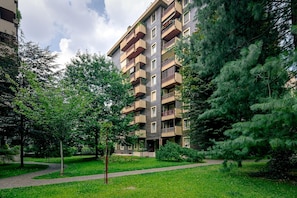 Um edifício residencial com vários andares e varandas, cercado por um jardim bem cuidado com árvores e um caminho pavimentado.