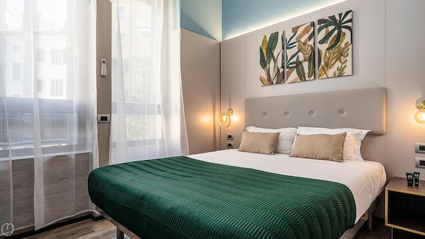 Una habitación de hotel con una cama grande, una colcha verde a rayas, una obra de arte colgada en la pared y una ventana con cortinas translúcidas.