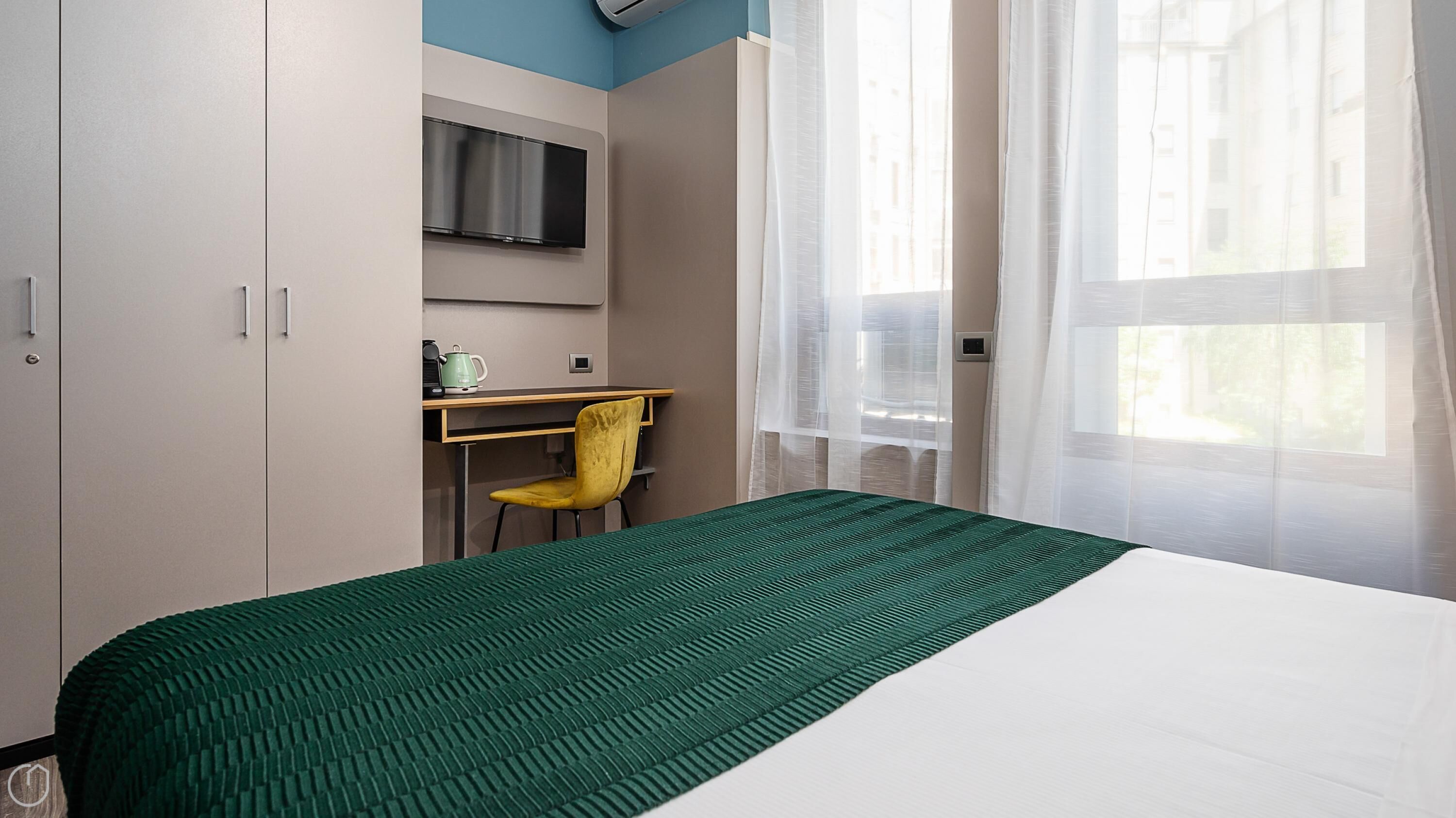 A hotel room with a bed, a desk with a chair, a television, and a window with sheer curtains.
