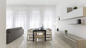 Una sala de estar moderna y bien iluminada con un sofá gris, una mesa de comedor con sillas y una zona de cocina con encimera y armarios.