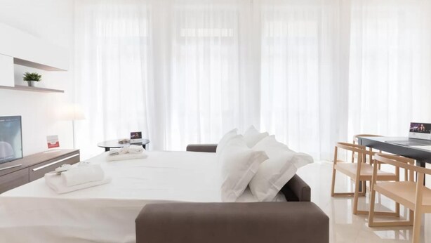 Habitación de hotel con cama, escritorio, silla, televisor y ventana con cortinas traslúcidas.