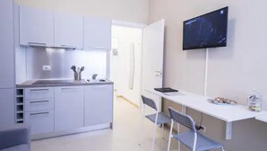 Una cocina moderna con armarios blancos, horno empotrado y lavamanos. Hay un escritorio blanco con una silla y un televisor de pantalla plana montado en la pared.