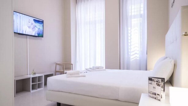 Habitación de hotel con cama, televisor montado en la pared, escritorio con silla y ventana con cortinas.