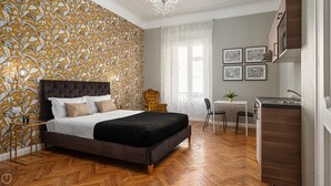 Une chambre avec un grand lit, un bureau et une chaise, une petite table avec une plante, et un coin cuisine avec un évier et des placards.