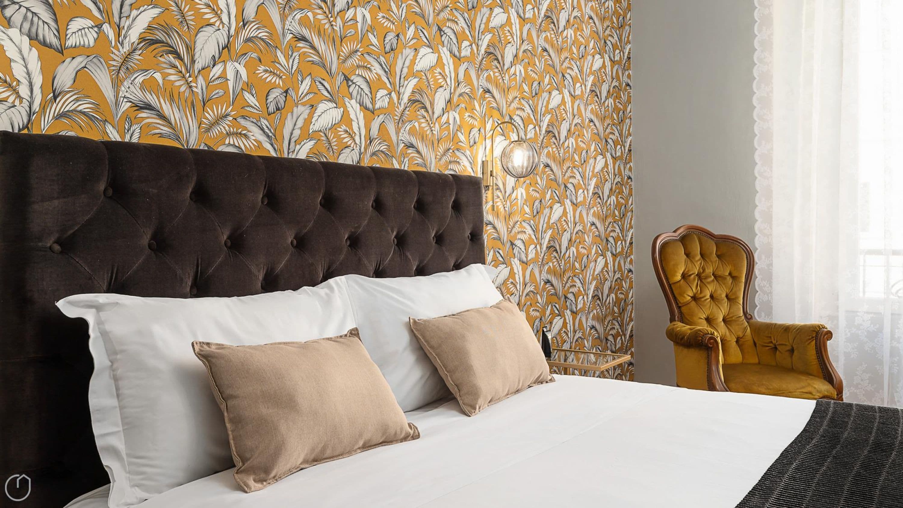 A bedroom with a tufted headboard, a bed with white and beige bedding, a yellow armchair, and a patterned wallpaper.