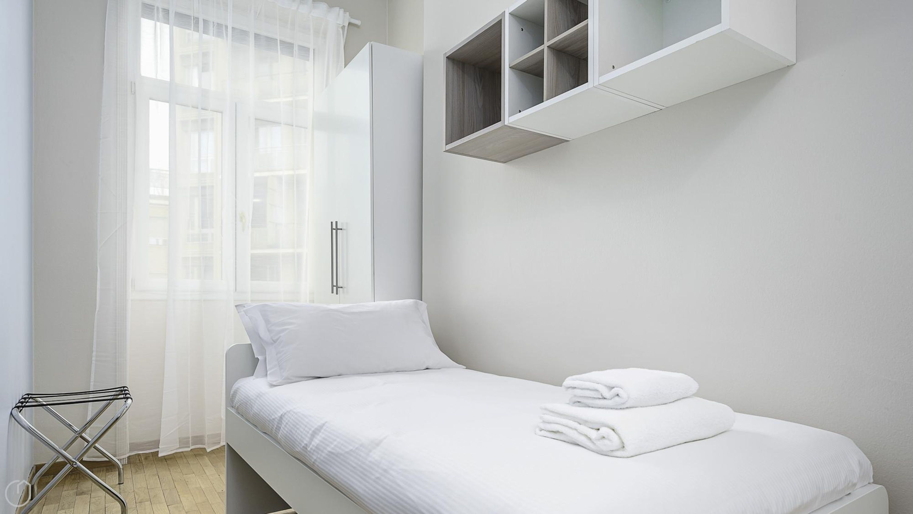 Een minimalistisch hotelkamer met een enkel bed, een klein nachtkastje en een raam met dunne gordijnen.
