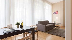 Un salon comprenant un canapé, une table à manger avec des chaises, une petite table d’appoint avec une lampe, et une grande fenêtre ornée de rideaux légers.