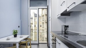 Une cuisine compacte avec un coin repas, une machine à café et une vue sur un immeuble d’habitation à travers une porte vitrée.