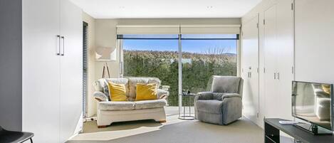 A modern living room with a sofa, armchair, and TV, large windows, and a view of the outdoors.