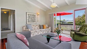 Un salon avec un canapé gris, un canapé à motifs floraux, un fauteuil vert et une table basse en verre. Un parapluie rouge se trouve à l’extérieur, près de la porte-fenêtre coulissante.
