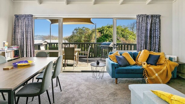 A spacious living area with a dining table, chairs, a blue sofa with cushions, and a view of a deck with a barbecue grill. - Mercy of Lake Taupo- family time + lake time (Taupo)