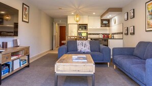 A modern living room with a sofa, a coffee table, and a bookshelf. There is a kitchen in the background with a microwave and a kettle. - Tui Unit - So close to the lake +Pool (Taupō)