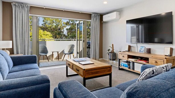 A modern living room with a sofa, a coffee table, a TV, and a balcony with a view of trees. - Tui Unit - So close to the lake +Pool (Taupō)