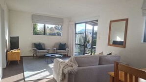 A spacious living room with a large sofa, a coffee table, and two armchairs. There is a television on a wooden stand, and a large window providing a view of the outdoors.