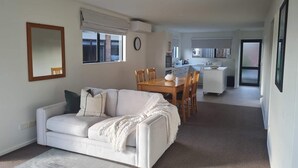 A living room with a white sofa, a dining table with chairs, and a kitchen in the background.