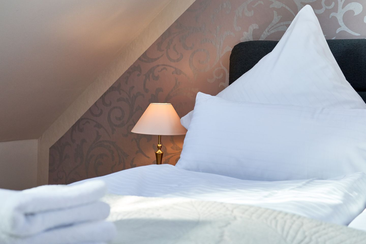 Een netjes opgemaakt bed met witte lakens en kussens, een bedlampje en behang met een patroon op de muur.