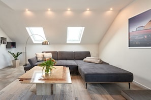 A modern loft bedroom with a large gray sectional sofa, a wooden coffee table, and a skylight.