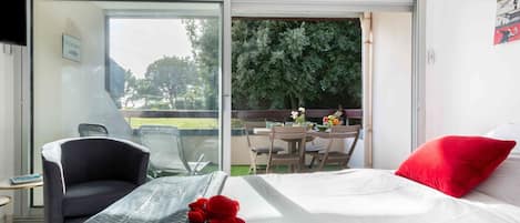 Et hotelværelse med en seng, en stol, et bord med en blomstervase og en balkon med udsigt til træer.