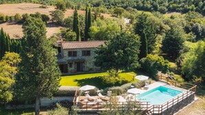 Una casa de piedra con piscina, rodeada de árboles y un césped cuidado.
