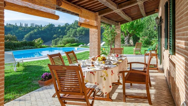 A covered outdoor dining area with a table set for breakfast, overlooking a swimming pool and lush greenery. - Villa Il Baiocco 8 in Ospedaletto (Urbino)