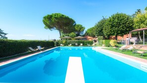 A rectangular swimming pool with a white lane divider, surrounded by lounge chairs and umbrellas, with a hedge and trees in the background.