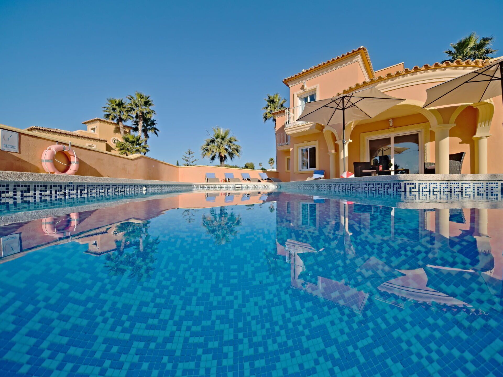A swimming pool with clear blue water, surrounded by a building with a tiled exterior and palm trees.