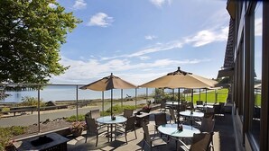 Een terras buiten met tafels en stoelen, grote parasols en uitzicht op een waterlichaam.