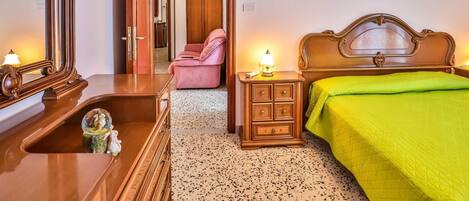 Un dormitorio con un aparador de madera, una cama con colcha verde, una mesita de noche con lámpara, una silla rosa y un armario de madera.