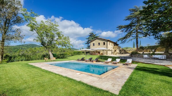 A stone house with a swimming pool, surrounded by a well-maintained lawn and trees.